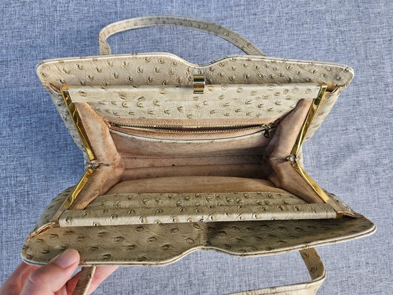 May include: A beige handbag with a textured ostrich-style pattern, featuring a gold-tone metal frame, a single top handle, and a light tan interior lining. The bag includes a zippered pocket inside and a secure metal clasp closure at the top.