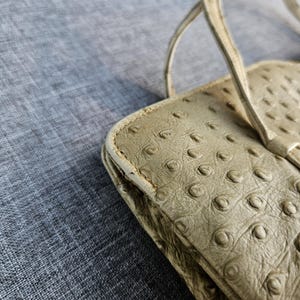 May include: A light beige handbag featuring a textured surface with a repeating pattern of raised circular bumps. The bag has thin, matching beige straps attached to the top edge, which is designed with a scalloped, wavy silhouette.