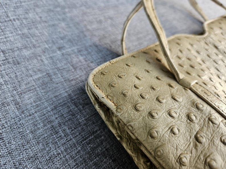 May include: A light beige handbag featuring a textured surface with a repeating pattern of raised circular bumps. The bag has thin, matching beige straps attached to the top edge, which is designed with a scalloped, wavy silhouette.