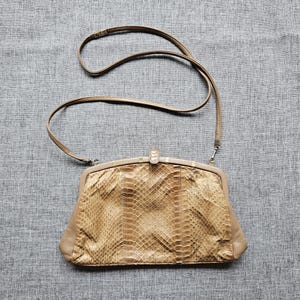 Può includere: Una borsa a mano color beige, in stile vintage, con un motivo a pelle di serpente sul davanti. La borsa ha una tracolla lunga e sottile e una chiusura a scatto. È in pelle e di forma rettangolare.