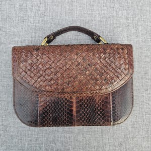 Könnte beinhalten: Eine braune Handtasche mit gewebter Textur auf der oberen Klappe und einem glatten Schuppenmuster am unteren Körper. Die Tasche verfügt über einen einzelnen Tragegriff, der mit goldfarbenen Metallbeschlägen befestigt ist und ein strukturiertes Aussehen verleiht.
