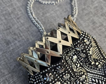 May include: A black handbag featuring a silver-toned metal accordion-style frame at the top and a silver chain handle. The exterior is covered in a dense pattern of small silver studs and flower-shaped metal embellishments across the fabric.