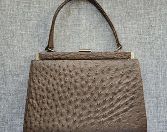 May include: A brown handbag with a textured, quill-patterned surface. The bag features a single top handle, a rectangular frame, and a silver-toned metal clasp closure at the top center. The design is structured with a flat base and tapered sides.