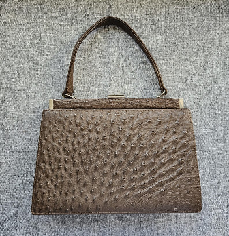 May include: A brown handbag with a textured, quill-patterned surface. The bag features a single top handle, a rectangular frame, and a silver-toned metal clasp closure at the top center. The design is structured with a flat base and tapered sides.