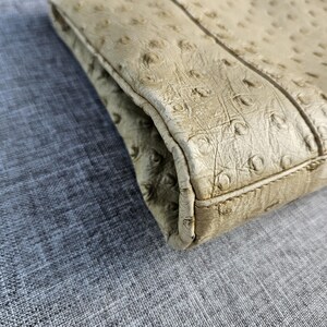 May include: A beige clutch with a distinct ostrich leather texture featuring a pattern of small, raised quill follicles across the surface. The accessory has a structured rectangular shape with visible stitched seams along the edges and a fold-over flap.