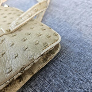 May include: A beige handbag featuring a textured surface with a repeating pattern of raised circular bumps. The bag has a structured rectangular edge with visible stitching along the seams and a thin handle attached to the top section.
