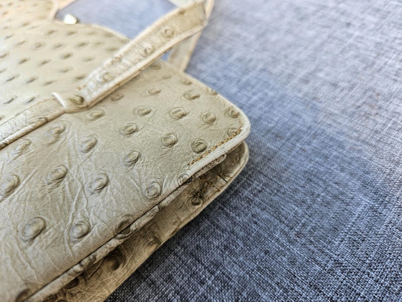 May include: A beige handbag featuring a textured surface with a repeating pattern of raised circular bumps. The bag has a structured rectangular edge with visible stitching along the seams and a thin handle attached to the top section.