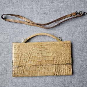 May include: A tan-coloured handbag with a crocodile skin pattern, featuring a top handle and a detachable shoulder strap. The bag has a rectangular shape with a flap closure and metal hardware.