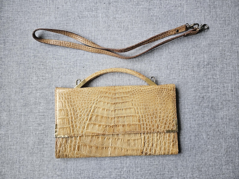May include: A tan-coloured handbag with a crocodile skin pattern, featuring a top handle and a detachable shoulder strap. The bag has a rectangular shape with a flap closure and metal hardware.