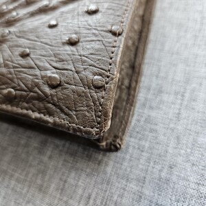 May include: A taupe-colored wallet featuring a textured surface with raised circular bumps characteristic of ostrich leather. The item has visible stitching along the edge, showcasing a structured corner design with a matte finish and neutral tone.