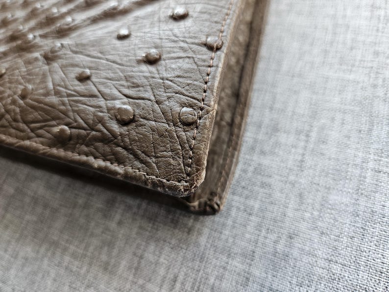 May include: A taupe-colored wallet featuring a textured surface with raised circular bumps characteristic of ostrich leather. The item has visible stitching along the edge, showcasing a structured corner design with a matte finish and neutral tone.