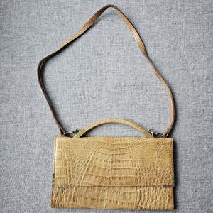 May include: A rectangular, light brown handbag with a crocodile-effect texture. It has a short handle and a long, thin strap. The bag's hardware is silver-toned, and it closes with a flap.