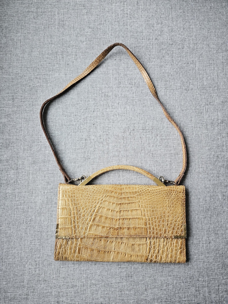 May include: A rectangular, light brown handbag with a crocodile-effect texture. It has a short handle and a long, thin strap. The bag's hardware is silver-toned, and it closes with a flap.