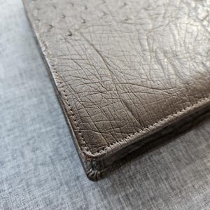 May include: A rectangular brown wallet featuring a textured ostrich leather pattern with visible quill follicles. The wallet has reinforced stitched edges and a metallic silver-toned clasp visible at the top corner of the folded flap closure.