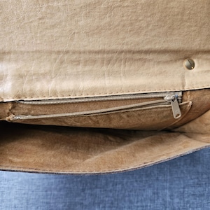 May include: An open tan leather clutch bag, showing the interior. It has a zipped pocket and a smaller open pocket. The bag is secured with a snap and a zip. The leather has a textured finish.