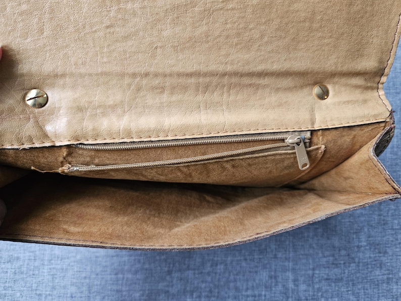 May include: An open tan leather clutch bag, showing the interior. It has a zipped pocket and a smaller open pocket. The bag is secured with a snap and a zip. The leather has a textured finish.