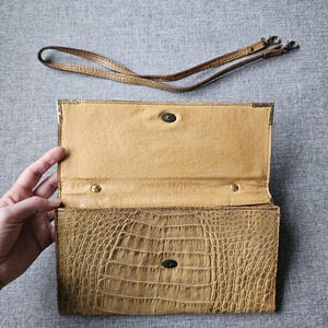 May include: Open, tan clutch bag with a crocodile-like texture. The interior is a lighter tan. A detachable, thin, brown strap lies above the open bag. The bag has a metal clasp and corner accents. The background is a grey fabric.