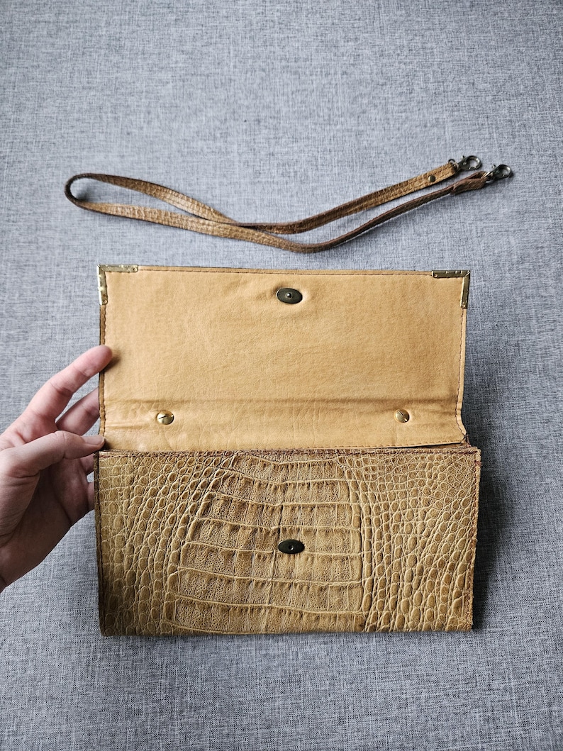 May include: Open, tan clutch bag with a crocodile-like texture. The interior is a lighter tan. A detachable, thin, brown strap lies above the open bag. The bag has a metal clasp and corner accents. The background is a grey fabric.