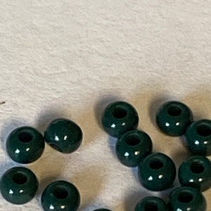 May include: A collection of dark green, round beads with a hole through the center. The beads are scattered on a light-colored surface. These beads are suitable for jewelry making and crafting projects.