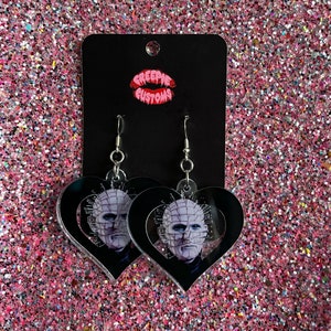 Może przedstawiać: Para czarnych kolczyków w kształcie serca z srebrnym haczykiem. Kolczyki przedstawiają grafikę postaci z filmu grozy 'Hellraiser'.