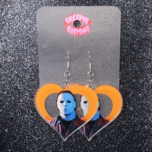 May include: A pair of orange heart-shaped earrings with a picture of Michael Myers from the Halloween movie franchise. The earrings are on a white card with the text "Creepie Customs".