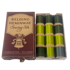 May include: A vintage Belding Heminway Sewing Silk box containing twelve wooden spools of thread in various shades of green. The brown box features an eagle logo, the text "BELDING HEMINWAY Sewing Silk", "FOUNDED 1849", and "WARRANTED 50 YARDS".
