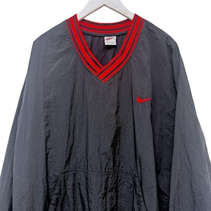 May include: A charcoal gray long-sleeve pullover windbreaker featuring a V-neckline with red and black striped ribbed trim. The garment includes a small red embroidered Nike swoosh logo on the left chest and a front kangaroo pocket.