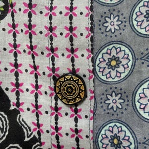 Pu&ograve; includere: Primo piano di tessuto fantasia con un bottone nero e oro. Il tessuto presenta un motivo floreale rosa e nero su sfondo grigio chiaro. Il bottone &egrave; rotondo con un motivo dorato dettagliato.