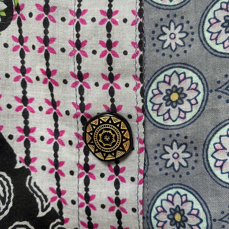 Pu&ograve; includere: Primo piano di tessuto fantasia con un bottone nero e oro. Il tessuto presenta un motivo floreale rosa e nero su sfondo grigio chiaro. Il bottone &egrave; rotondo con un motivo dorato dettagliato.