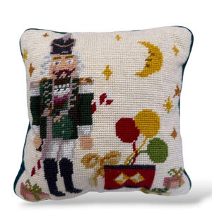May include: A square needlepoint pillow featuring a nutcracker in a green uniform standing next to a red box with three balloons in red, yellow, and green. The cream-colored background includes a yellow crescent moon and several small stars.