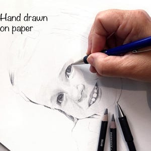 May include: A hand drawing a portrait of a child with a pencil on white paper. The text "Hand drawn on paper" is written in the top left corner of the page.