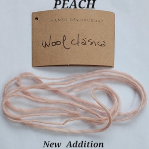 May include: A light peach colored yarn strand positioned below a brown paper tag. The tag features the printed text PEACH, MANOS DEL URUGUAY, and the handwritten text Wool clasica, with the words New Addition printed at the bottom of the image.