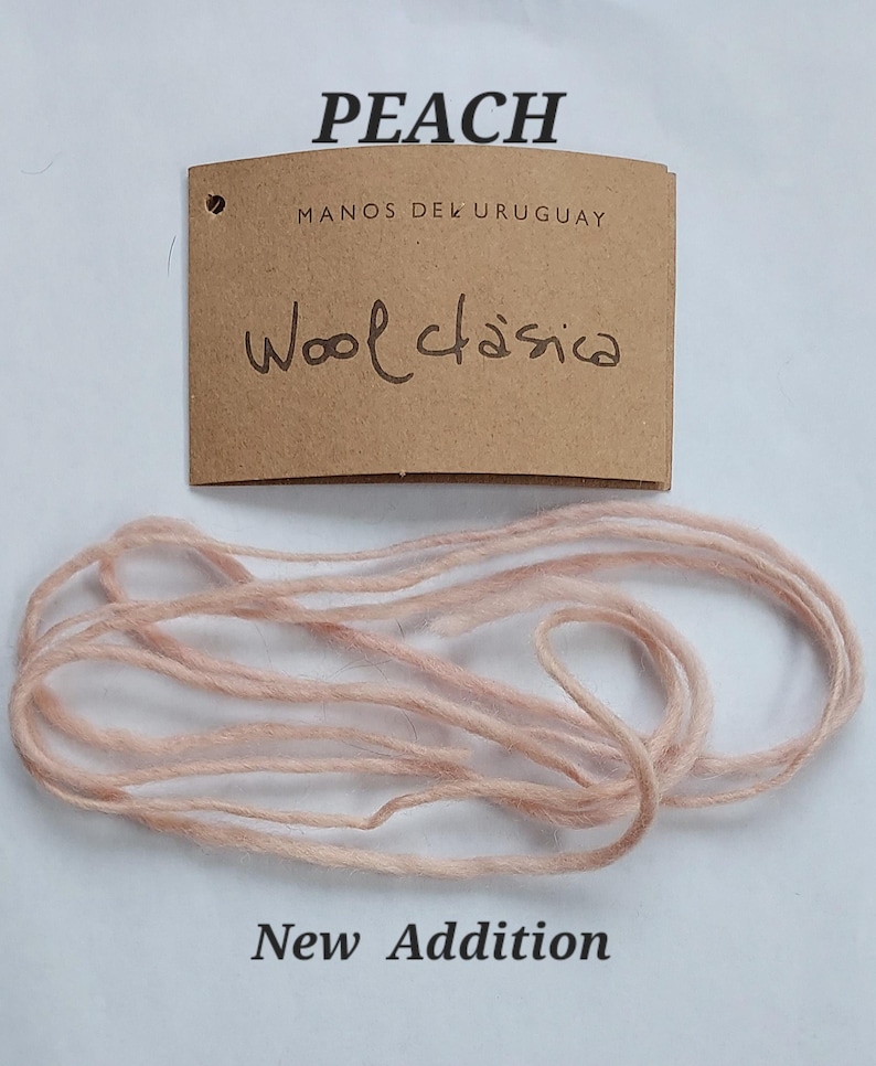 May include: A light peach colored yarn strand positioned below a brown paper tag. The tag features the printed text PEACH, MANOS DEL URUGUAY, and the handwritten text Wool clasica, with the words New Addition printed at the bottom of the image.