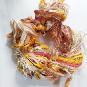 May include: A circular bundle of assorted decorative fibers and ribbons in shades of yellow, peach, pink, and metallic gold. The texture includes thick twisted yarn, frayed fabric strips, and shimmering gold-threaded ribbons woven together.