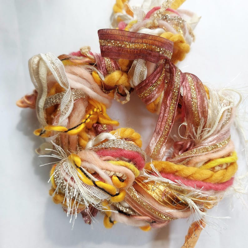 May include: A circular bundle of assorted decorative fibers and ribbons in shades of yellow, peach, pink, and metallic gold. The texture includes thick twisted yarn, frayed fabric strips, and shimmering gold-threaded ribbons woven together.