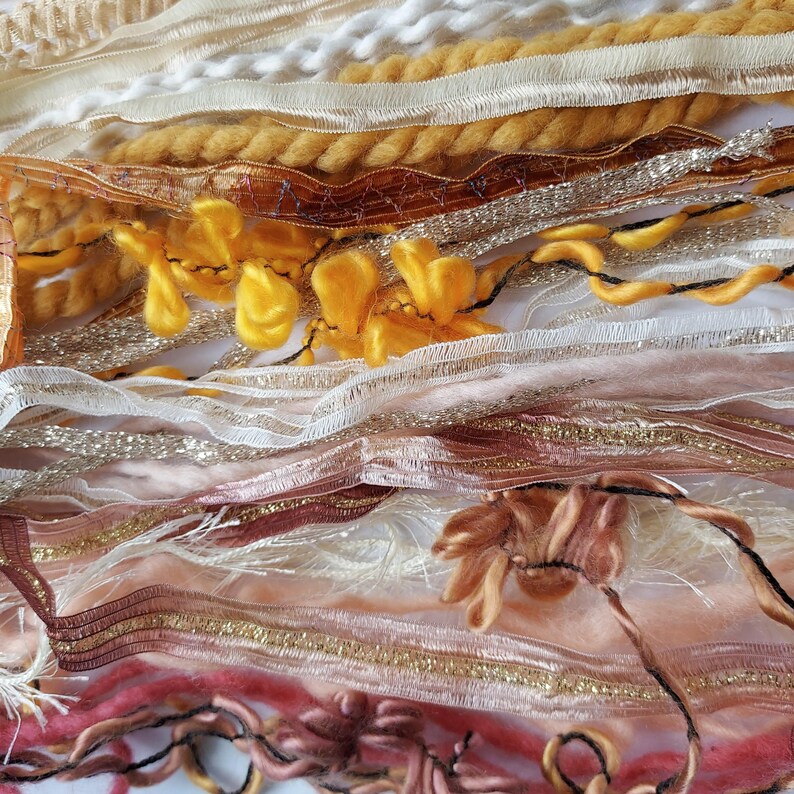 May include: A collection of assorted decorative ribbons and yarns in shades of gold, cream, copper, and soft pink. The assortment includes textured metallic threads, thick twisted wool, sheer ruffled trim, and delicate fringed fabric strips.