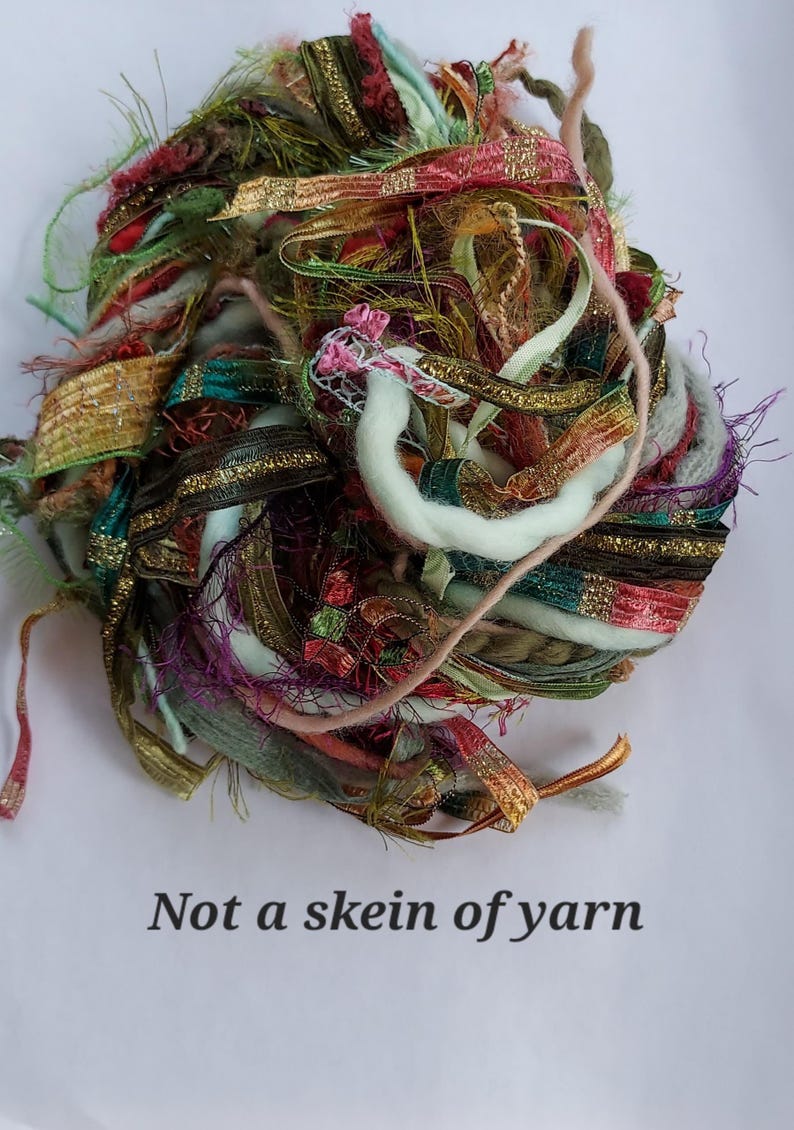 May include: A decorative bundle of assorted textured ribbons and fibers in shades of green, red, gold, brown, and pink, featuring metallic accents and frayed edges. The text "Not a skein of yarn" is printed in black below the fiber collection.