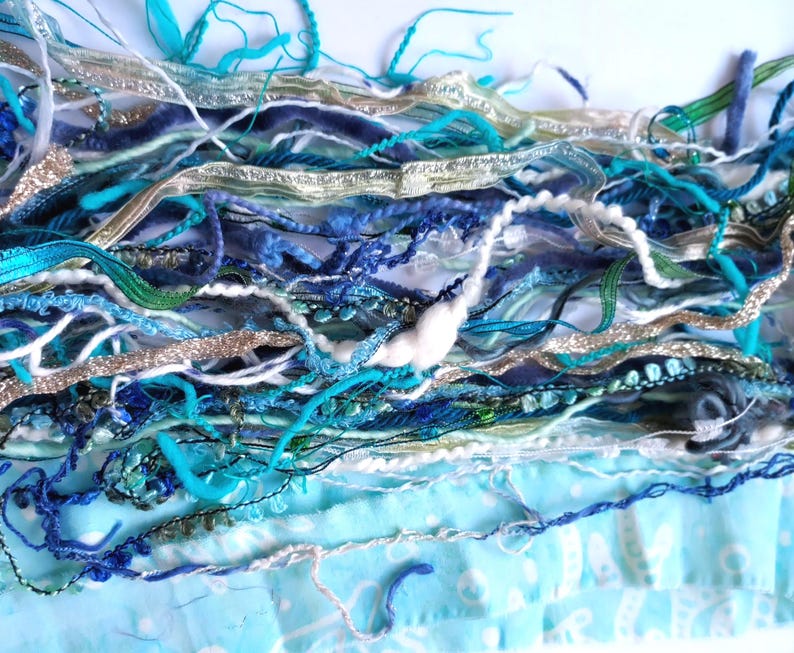 May include: A collection of assorted decorative ribbons and yarns in shades of turquoise, navy blue, light blue, white, and metallic silver. The bundle includes various textures such as velvet, braided cord, shimmering metallic strips, and soft wool.