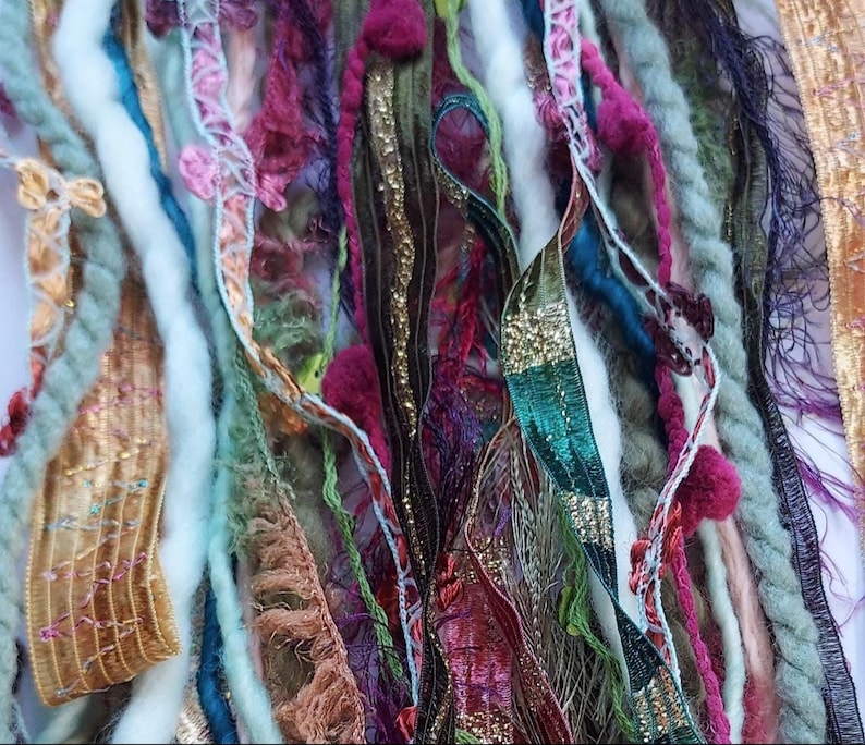 May include: A collection of assorted decorative ribbons and yarn strands in various textures and colors, including gold, magenta, teal, sage green, and white. The assortment features metallic threads, pom-poms, frayed edges, and woven patterns.