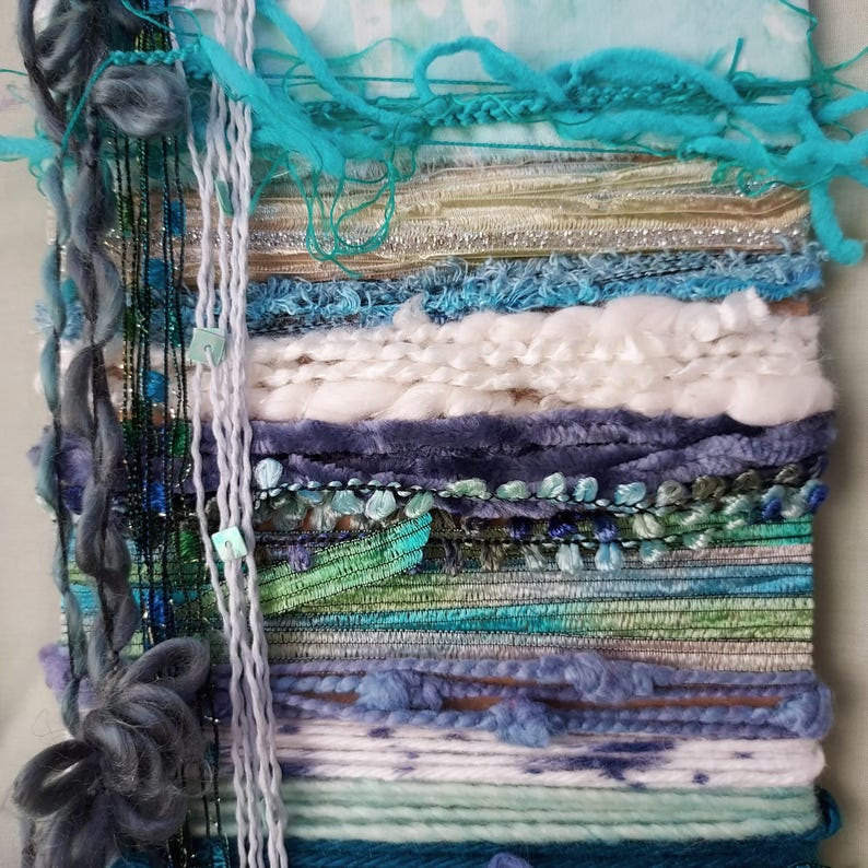 May include: A wall hanging tapestry featuring a layered composition of various fibers, including turquoise, white, navy, and green yarn, velvet ribbons, metallic threads, and braided cords, creating a textured, multi-dimensional surface.