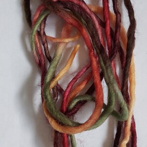May include: A coiled length of kettle-dyed wool yarn featuring variegated shades of dark brown, deep red, olive green, and golden yellow. The text NEW ADDITION appears at the top, and Manos del Uruguay kettle-dyed wool is at the bottom.