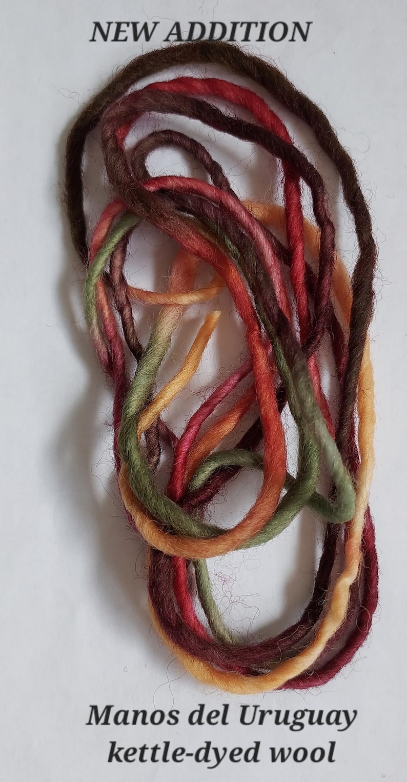 May include: A coiled length of kettle-dyed wool yarn featuring variegated shades of dark brown, deep red, olive green, and golden yellow. The text NEW ADDITION appears at the top, and Manos del Uruguay kettle-dyed wool is at the bottom.
