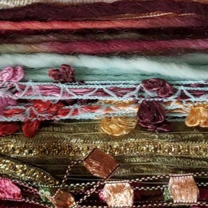 May include: A collection of assorted textile ribbons and yarns stacked horizontally. The assortment includes textured wool, metallic gold ribbon, delicate lace trim with pink and burgundy accents, and thick white roving yarn on the right side.