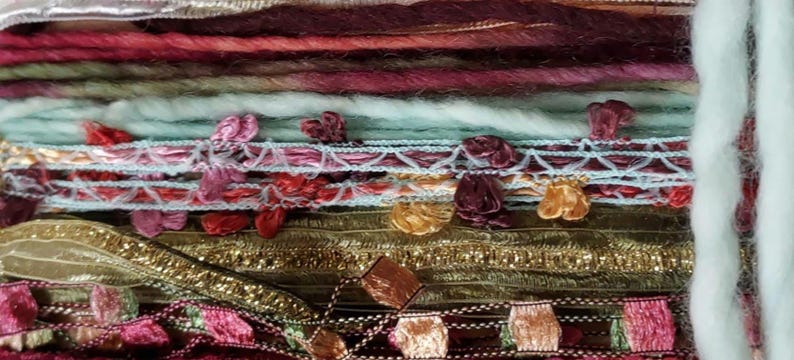 May include: A collection of assorted textile ribbons and yarns stacked horizontally. The assortment includes textured wool, metallic gold ribbon, delicate lace trim with pink and burgundy accents, and thick white roving yarn on the right side.