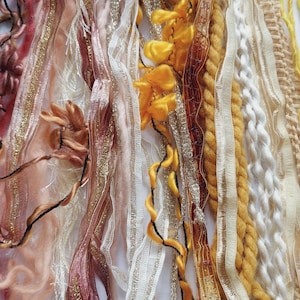 May include: A collection of assorted decorative ribbons and yarns in shades of yellow, gold, white, and dusty rose. The assortment includes metallic gold trim, twisted rope, ruffled fabric, sheer lace, and soft yarn strands arranged vertically.