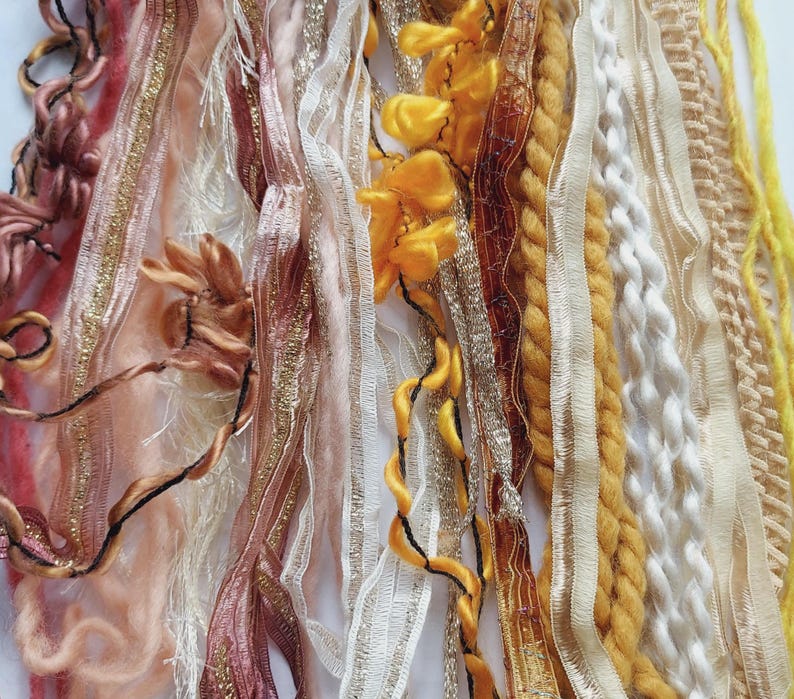 May include: A collection of assorted decorative ribbons and yarns in shades of yellow, gold, white, and dusty rose. The assortment includes metallic gold trim, twisted rope, ruffled fabric, sheer lace, and soft yarn strands arranged vertically.