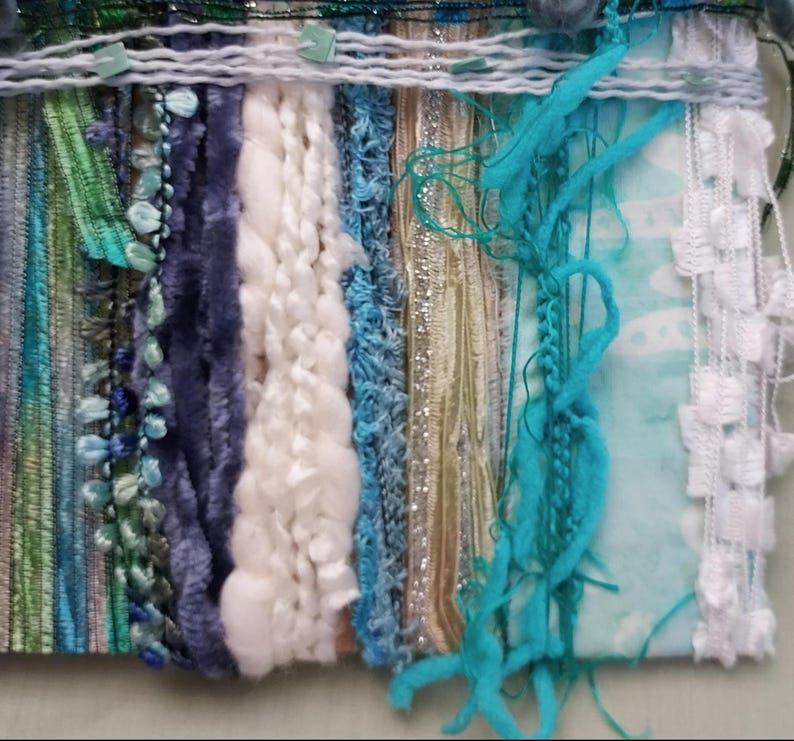 May include: A textile art piece featuring a vertical arrangement of varied fibers including white chunky yarn, deep purple velvet ribbon, shimmering metallic gold strips, turquoise fringe, and white lace trim, all woven together in a multi-textured display.