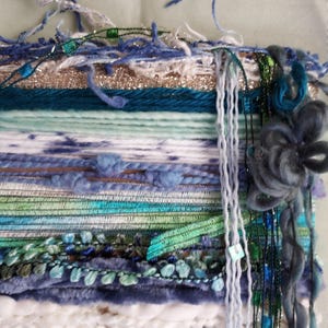 May include: A wall hanging tapestry featuring a horizontal arrangement of various yarn textures and fibers in shades of blue, green, white, and metallic gold. The piece includes braided elements, fuzzy yarn, and a large, dark blue fiber flower.