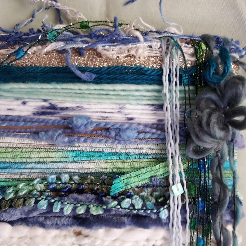 May include: A wall hanging tapestry featuring a horizontal arrangement of various yarn textures and fibers in shades of blue, green, white, and metallic gold. The piece includes braided elements, fuzzy yarn, and a large, dark blue fiber flower.