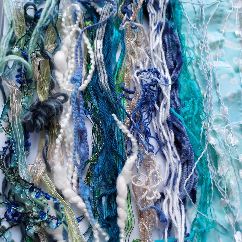 May include: A collection of assorted decorative ribbons and yarns in shades of blue, teal, white, and silver. The assortment includes textured fibers, metallic threads, velvet strips, and beaded strands arranged vertically in a dense, layered display.